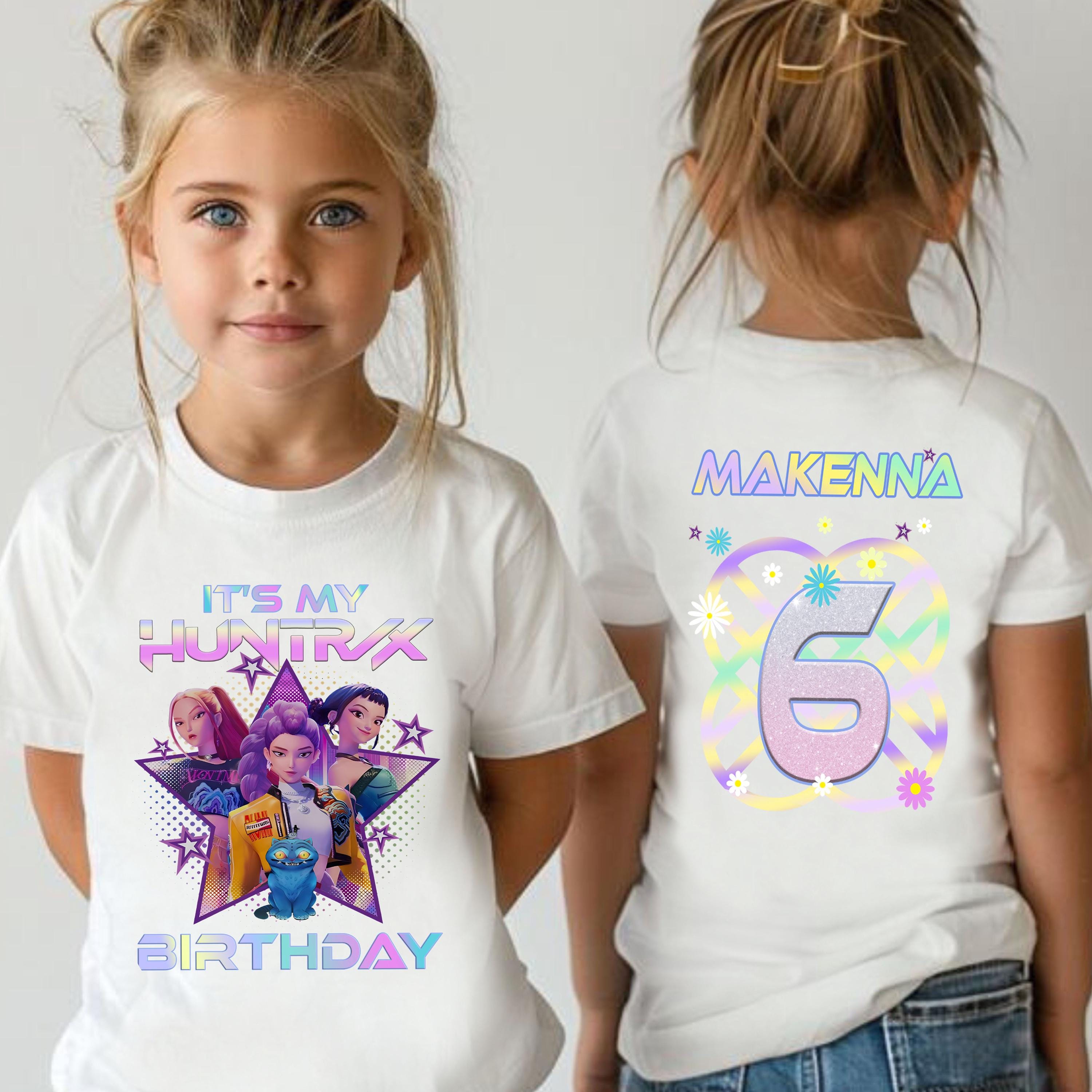 A young girl wearing a white t-shirt with a colorful graphic design and text, including the name 'MAKENNA' and the number '6'.