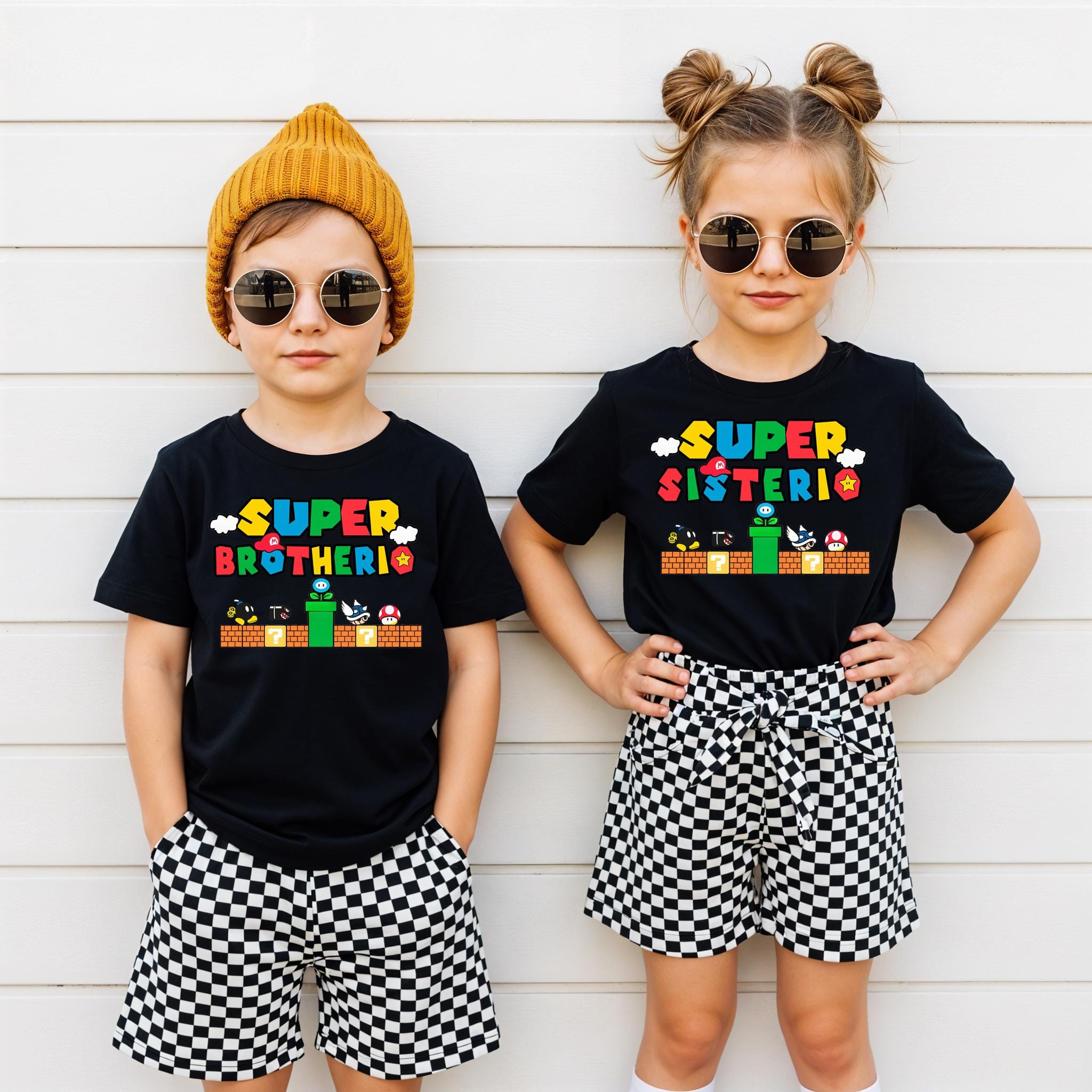 Two children wearing black t-shirts with a colorful design and sunglasses, standing against a white wall.