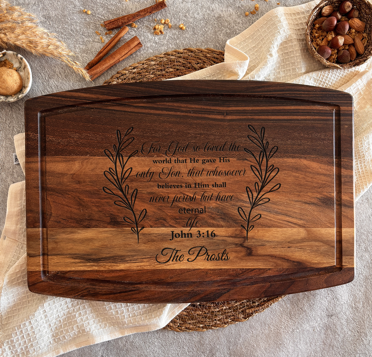 A wooden cutting board with a Bible verse engraved on it, surrounded by cinnamon sticks, a bowl of nuts, and a woven placemat.