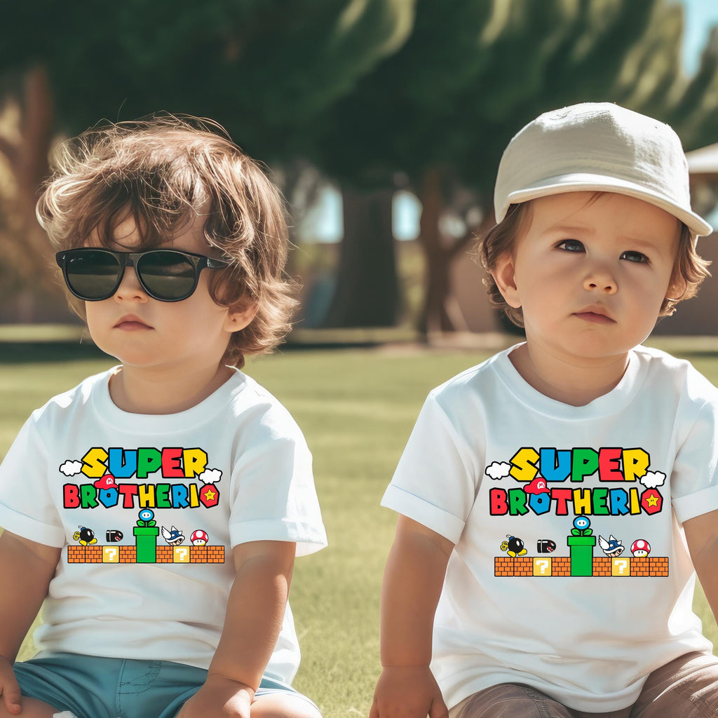 Two young children wearing white t-shirts with the text "SUPER BROTHERO" and a Mario-themed graphic, sitting on the grass.