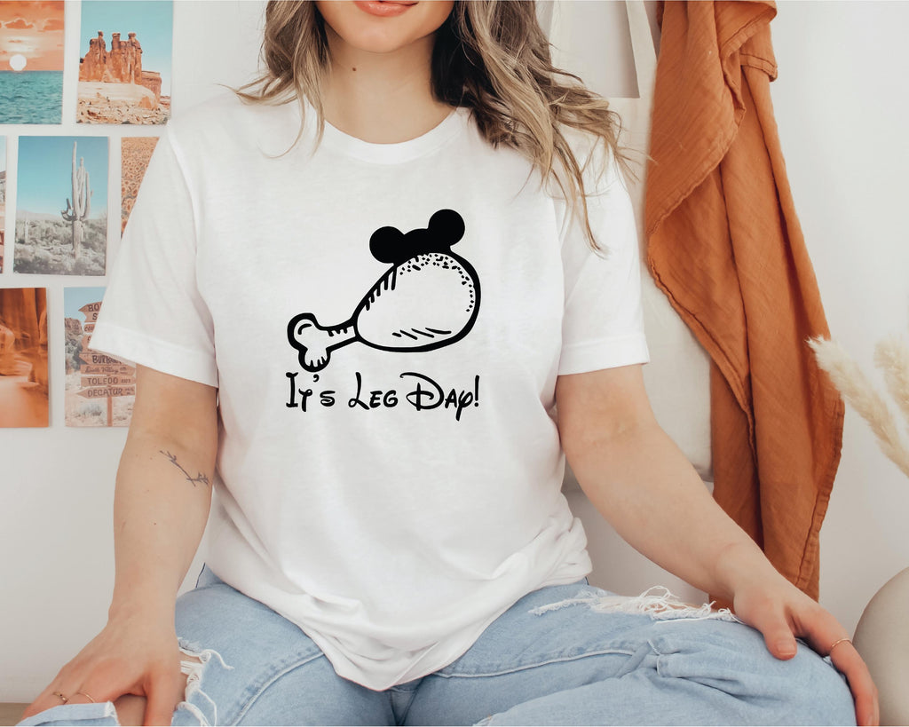 A woman wearing a white t-shirt with a graphic of a chicken leg and the text "It's Leg Day!" printed on it.
