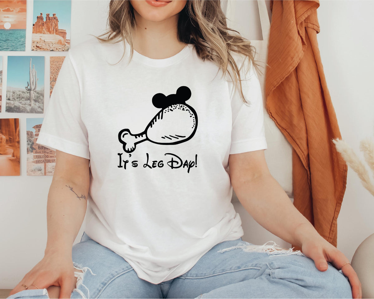 A woman wearing a white t-shirt with a graphic of a chicken leg and the text "It's Leg Day!" printed on it.