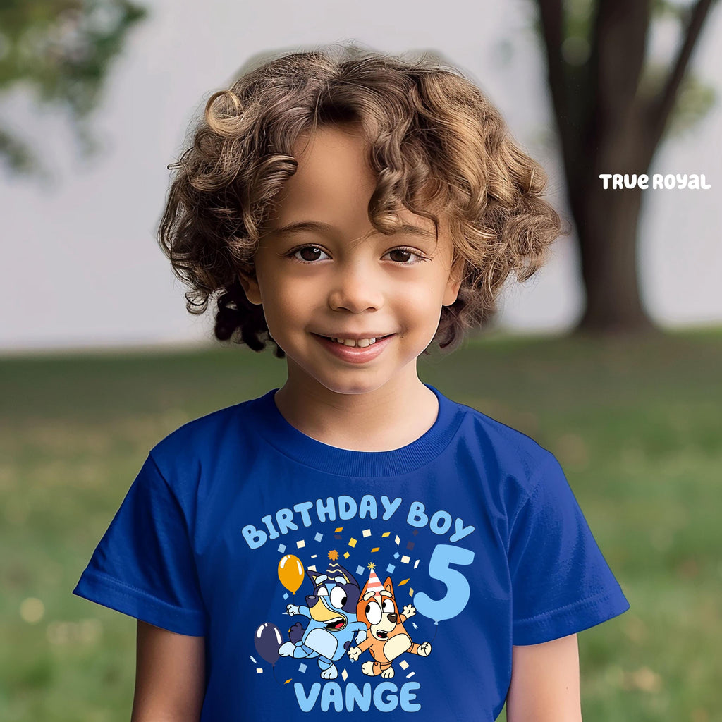 A young boy with curly hair wearing a blue t-shirt with a cartoon character and text, smiling at the camera.
