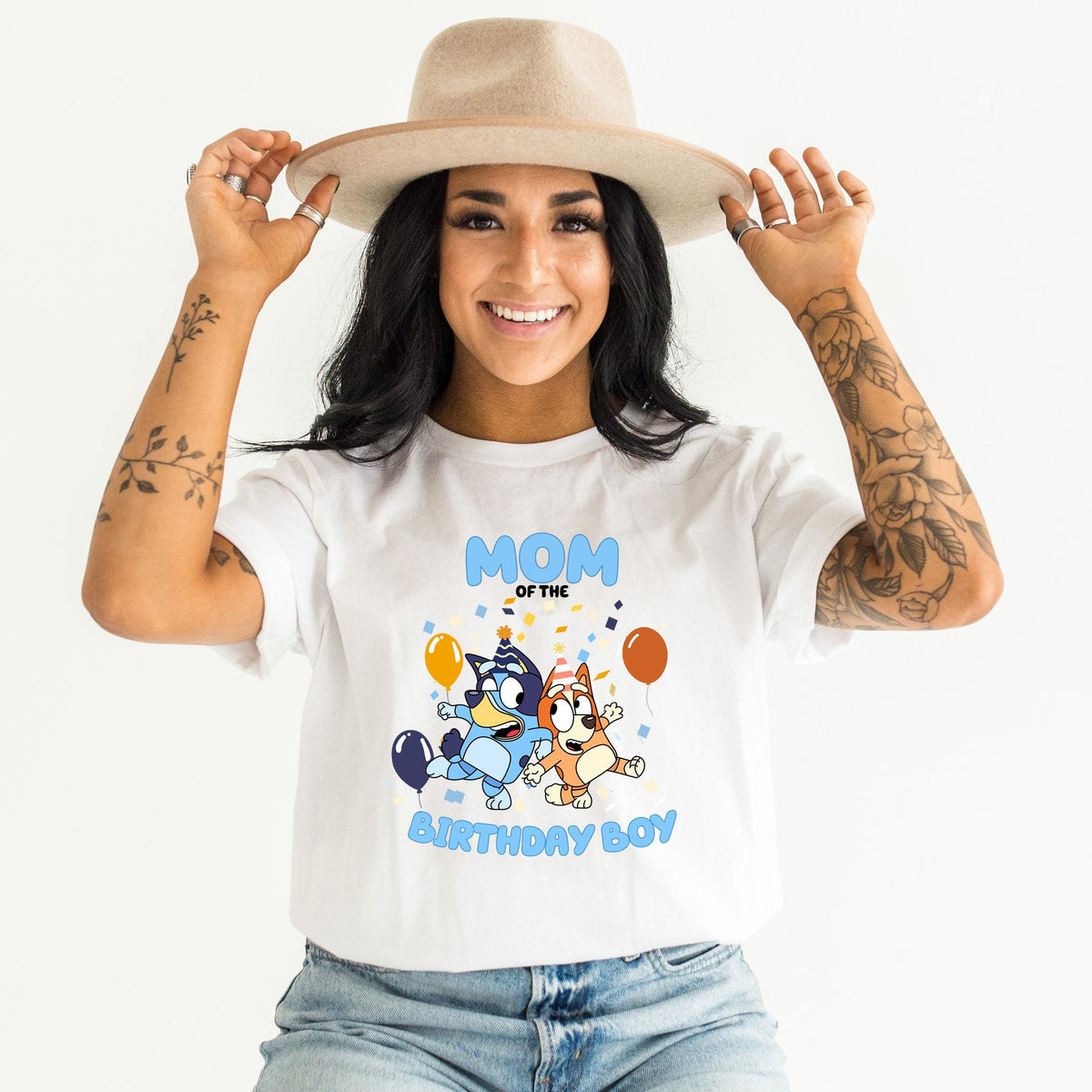 A woman wearing a white t-shirt with a graphic of two cartoon dogs and the text "MOM OF THE BIRTHDAY BOY" is smiling and adjusting her hat.
