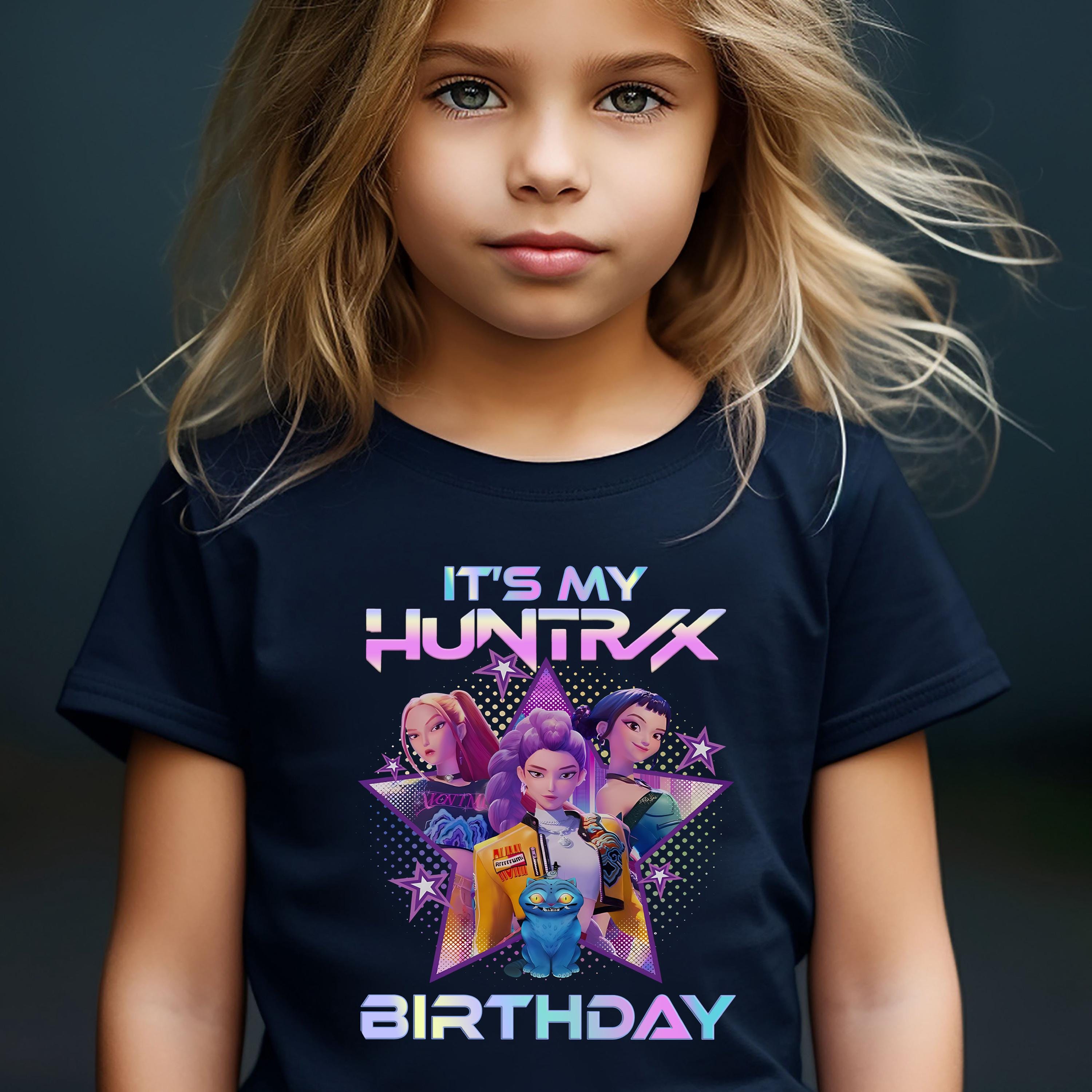 A young girl with blonde hair wearing a black t-shirt with a colorful graphic design featuring characters from the video game "It's My Huntx".
