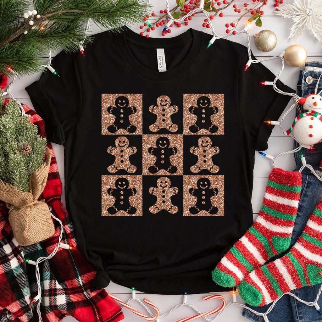 A black t-shirt with a graphic design of nine glittery gingerbread man figures arranged in a 3x3 grid on a white background. The t-shirt is surrounded by Christmas decorations, including a pine tree, ornaments, and Christmas lights.