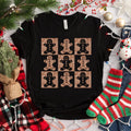 A black t-shirt with a graphic design of nine glittery gingerbread man figures arranged in a 3x3 grid on a white background. The t-shirt is surrounded by Christmas decorations, including a pine tree, ornaments, and Christmas lights.