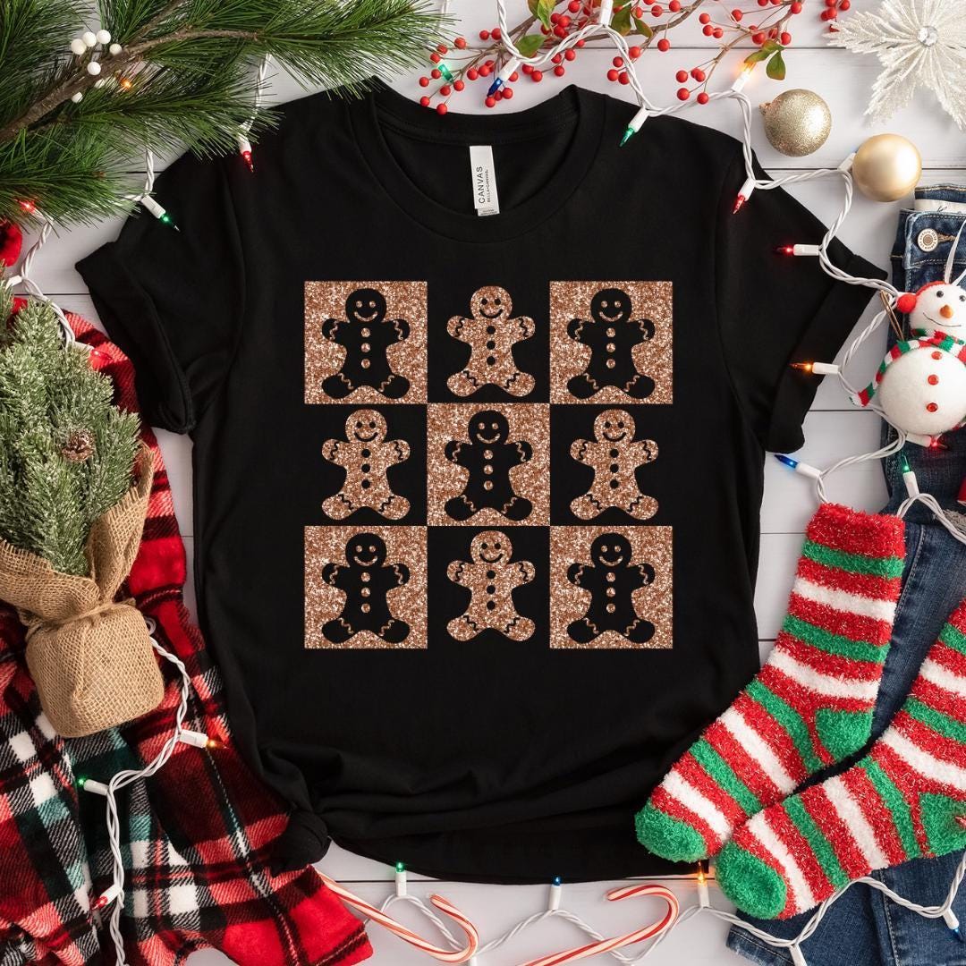 A black t-shirt with a graphic design of nine glittery gingerbread man figures arranged in a 3x3 grid on a white background. The t-shirt is surrounded by Christmas decorations, including a pine tree, ornaments, and Christmas lights.