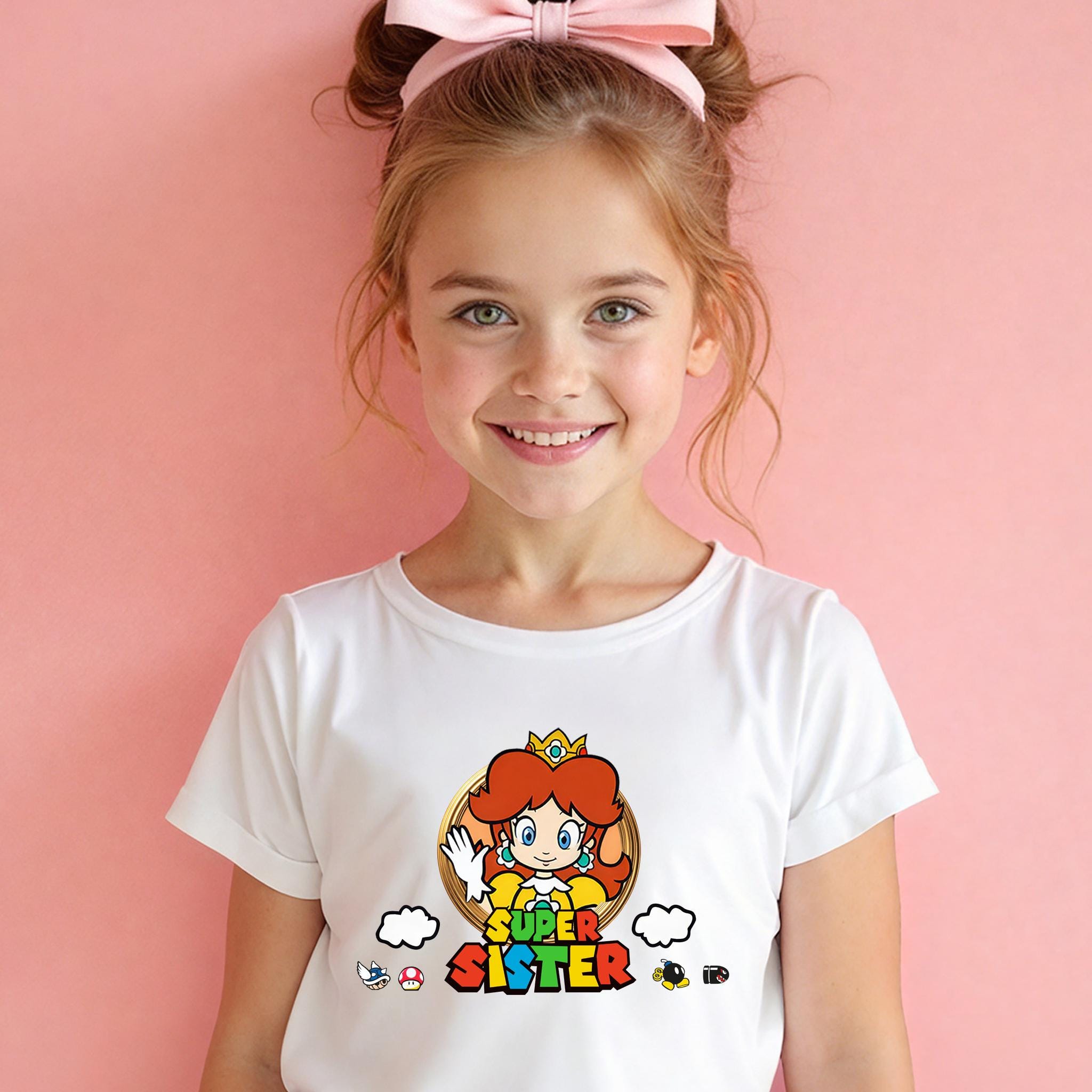 A young girl with a pink bow in her hair is smiling and wearing a white t-shirt with a colorful graphic of a character from the Super Mario video game series.