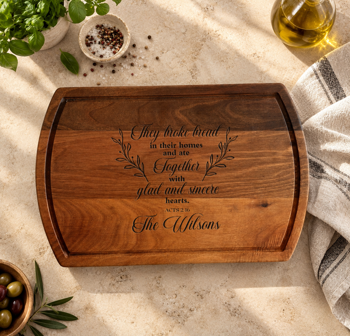 a wooden cutting board with a quote engraved on it, surrounded by various kitchen items such as a bowl of olives, a potted plant, and a glass of olive oil.