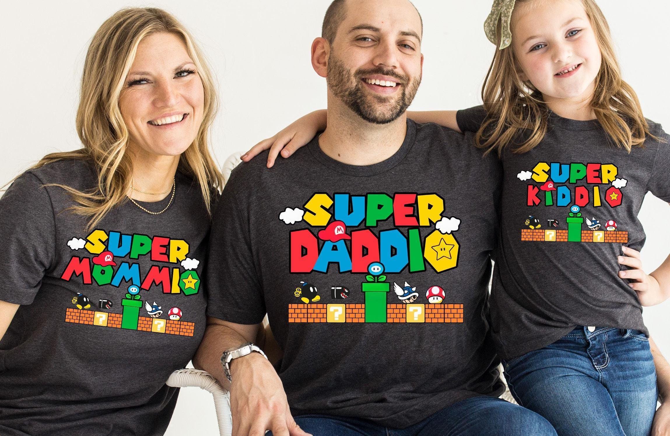 A man and two children are sitting together, smiling and wearing matching t-shirts with the text "SUPER KIDDIO" printed on them.