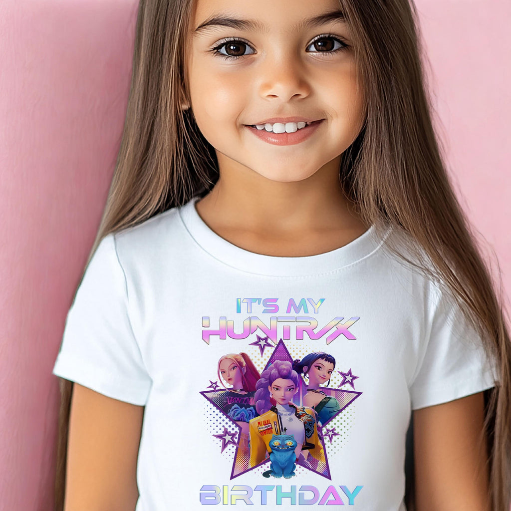 A young girl wearing a white t-shirt with a graphic design featuring three cartoon characters and the text "It's my HUNTRX birthday".