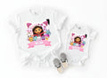 A white t-shirt with a cartoon image of a girl surrounded by various cartoon characters, and a smaller white baby onesie with the same cartoon image and the name "Amelia" printed on it.