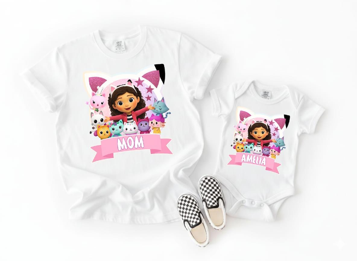 A white t-shirt with a cartoon image of a girl surrounded by various cartoon characters, and a smaller white baby onesie with the same cartoon image and the name "Amelia" printed on it.