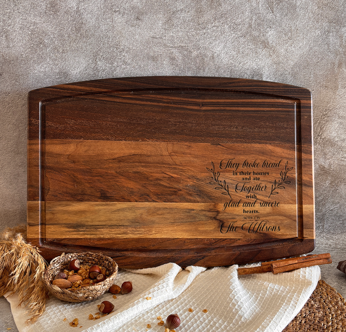 a wooden cutting board with a message engraved on it, placed on a woven mat and surrounded by various food items such as nuts and a bowl of fruit.