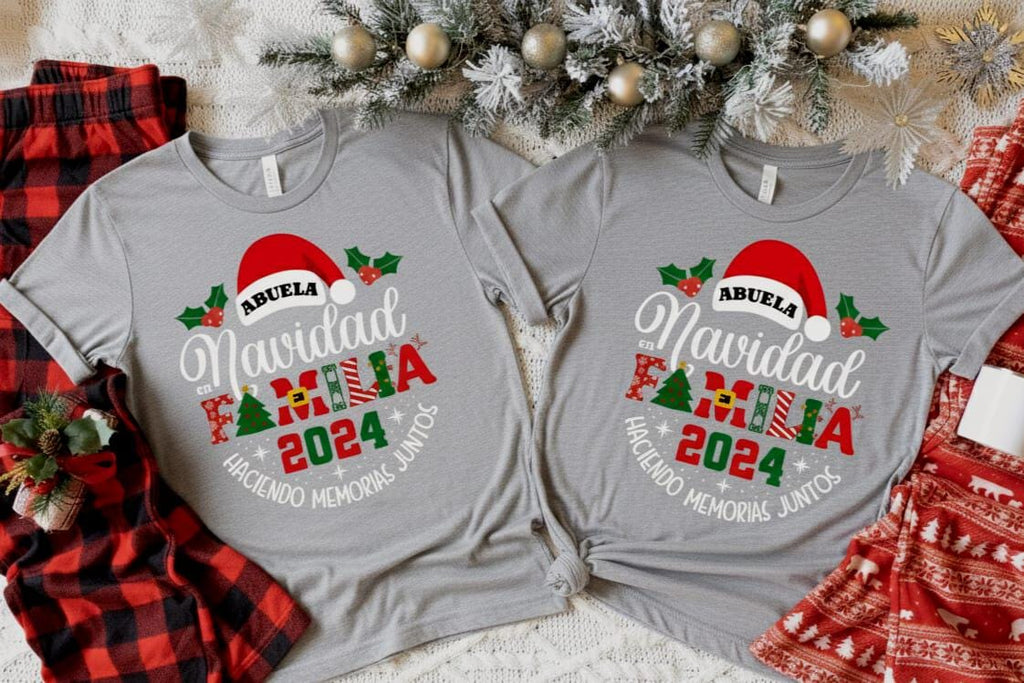 Two gray t-shirts with festive holiday-themed designs, including a Santa hat and Christmas trees, are displayed on a bed with a red and black plaid blanket and a Christmas tree.