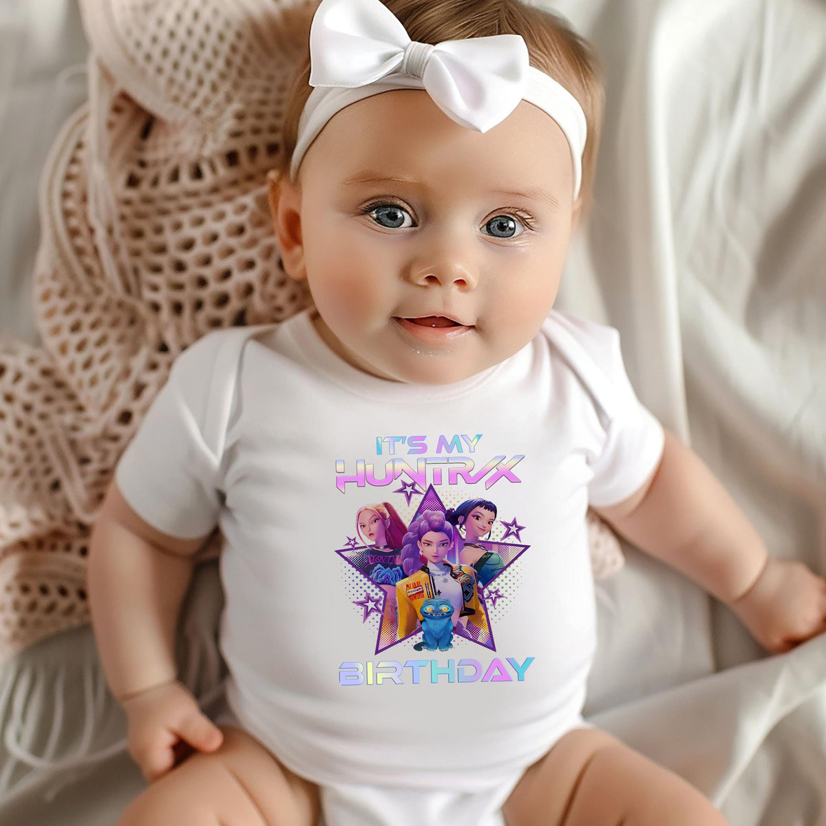 A smiling baby girl wearing a white onesie with a colorful graphic and text on it.