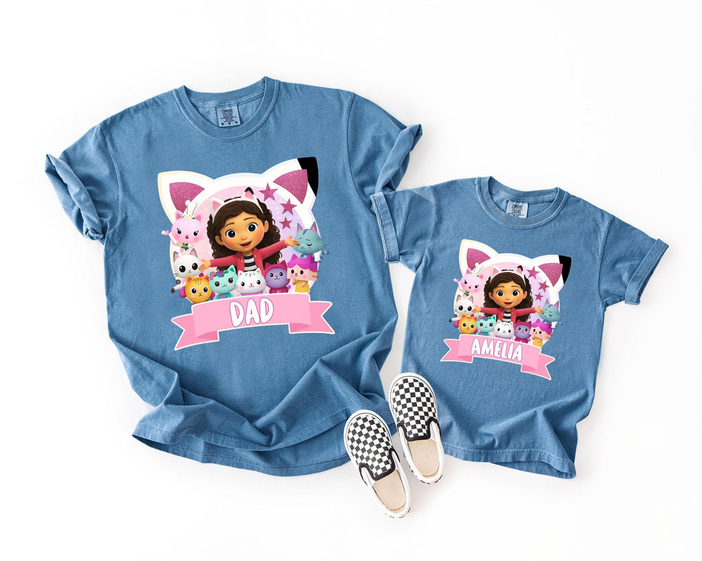 A blue t-shirt with a cartoon image of a girl and a cat, along with the text "DAD" and a smaller t-shirt with the same design and text.