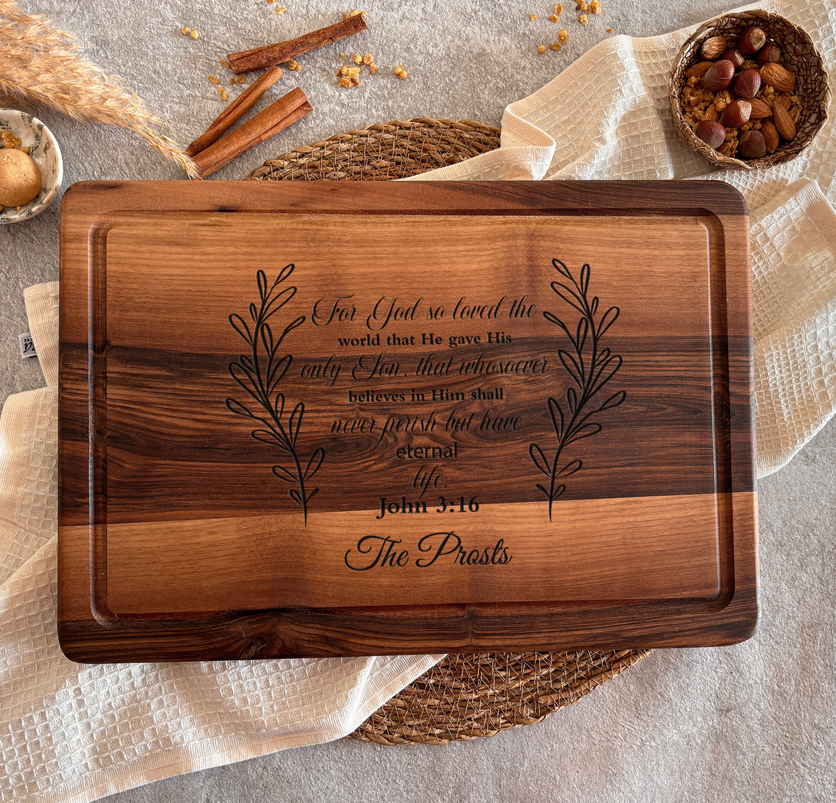 A wooden cutting board with a Bible verse engraved on it, surrounded by cinnamon sticks, a woven placemat, and a bowl of nuts.