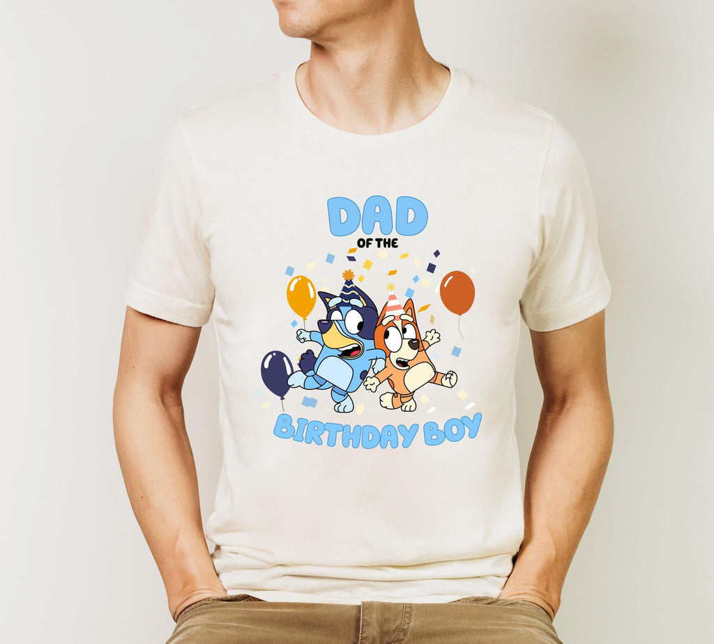 A white t-shirt with a graphic design featuring two cartoon dogs, one blue and one brown, along with balloons and confetti, along with the text "DAD OF THE BIRTHDAY BOY".