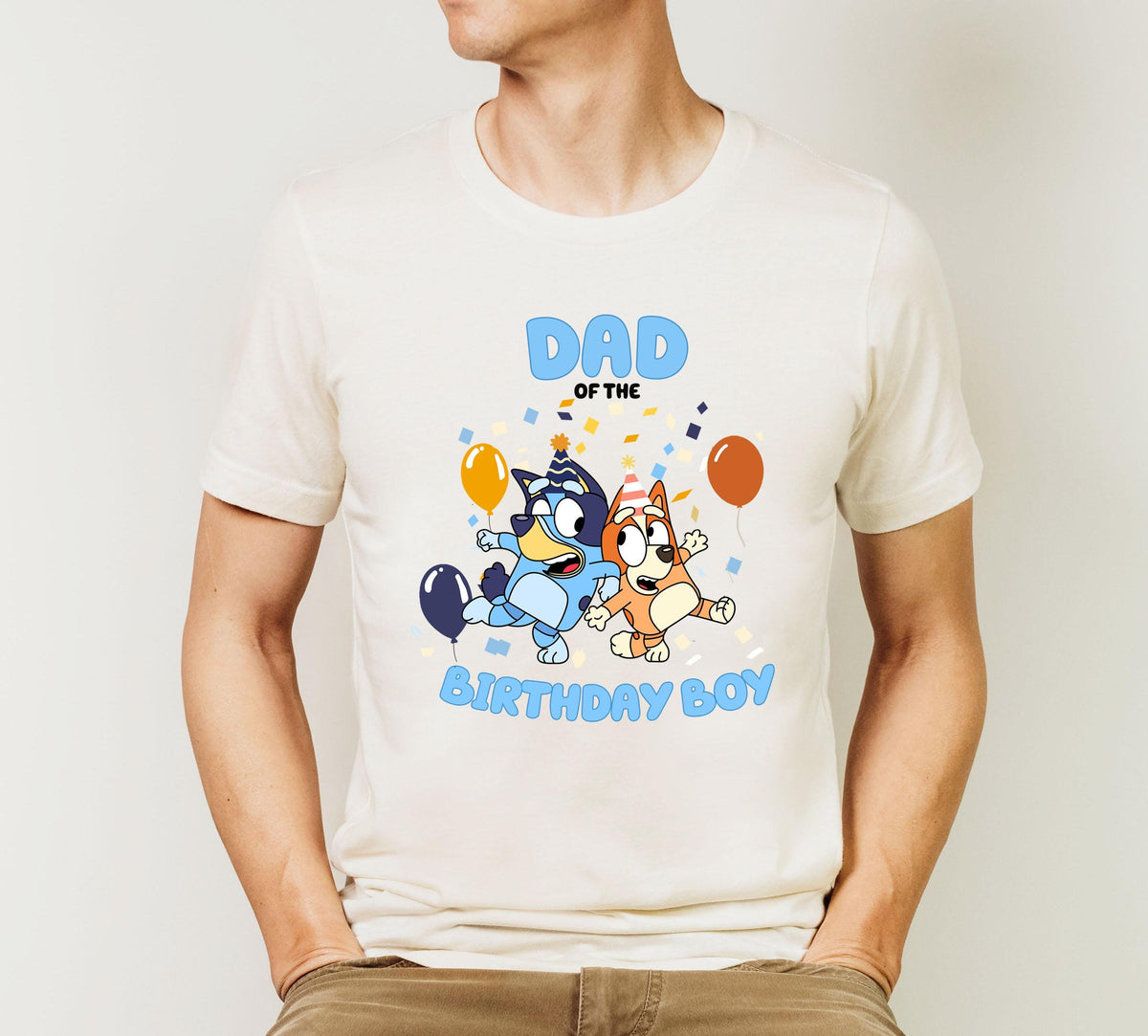 A white t-shirt with a graphic design featuring two cartoon dogs, one blue and one brown, along with balloons and confetti, along with the text "DAD OF THE BIRTHDAY BOY".